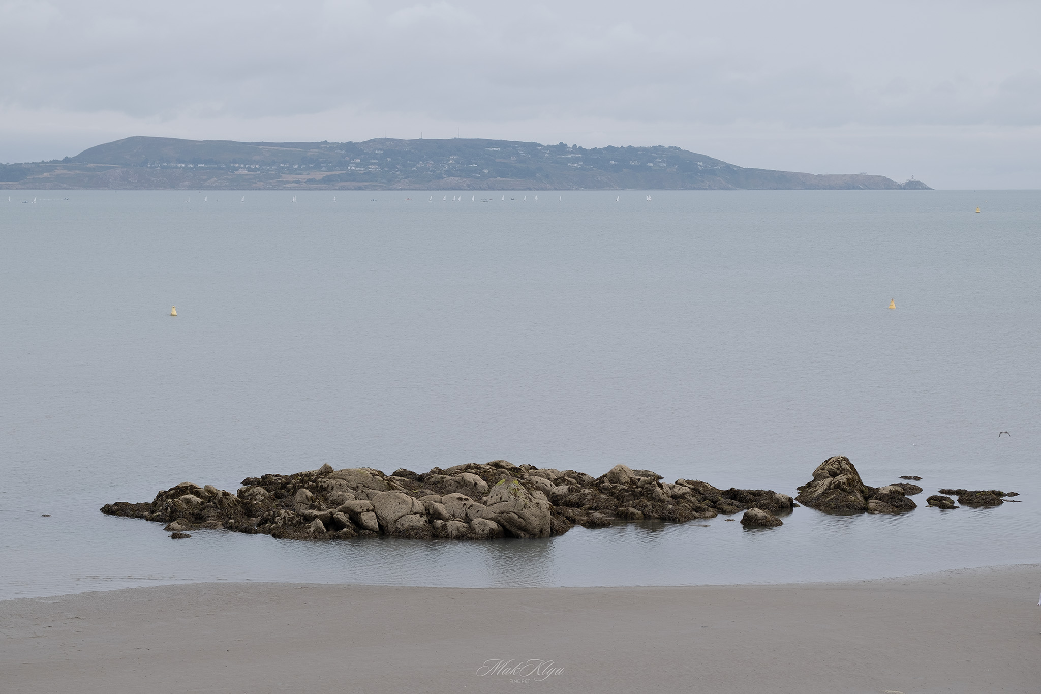 Rocky outcrop in a calm Irish bay with distant hills and sailboats under a cloudy sky