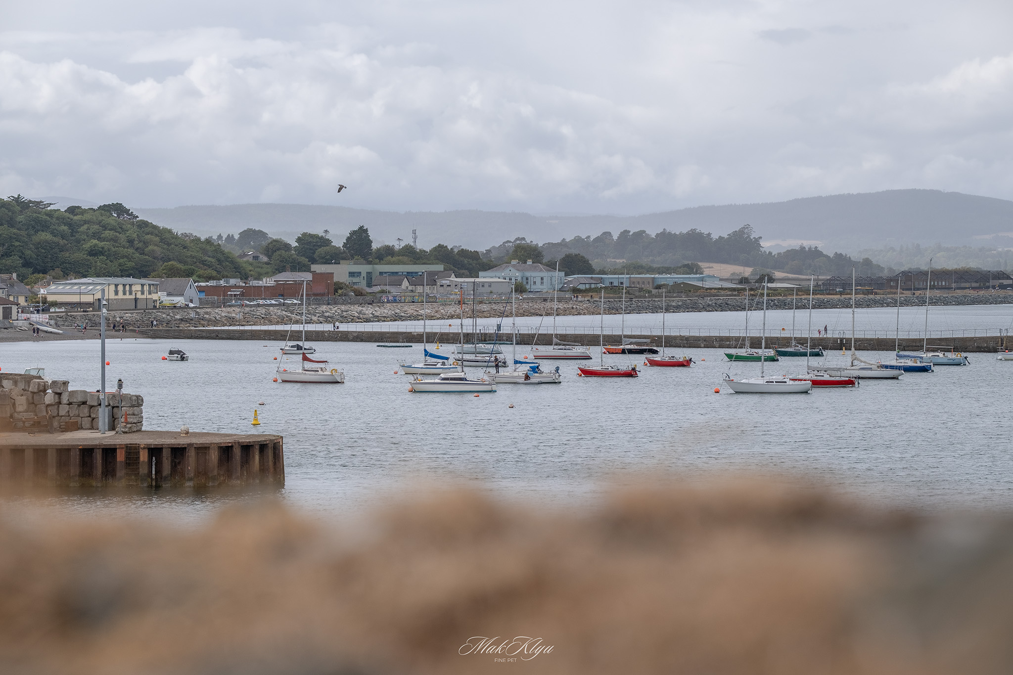 Small sailboats moored in a quiet coastal harbour beneath cloudy hills and a grey sky