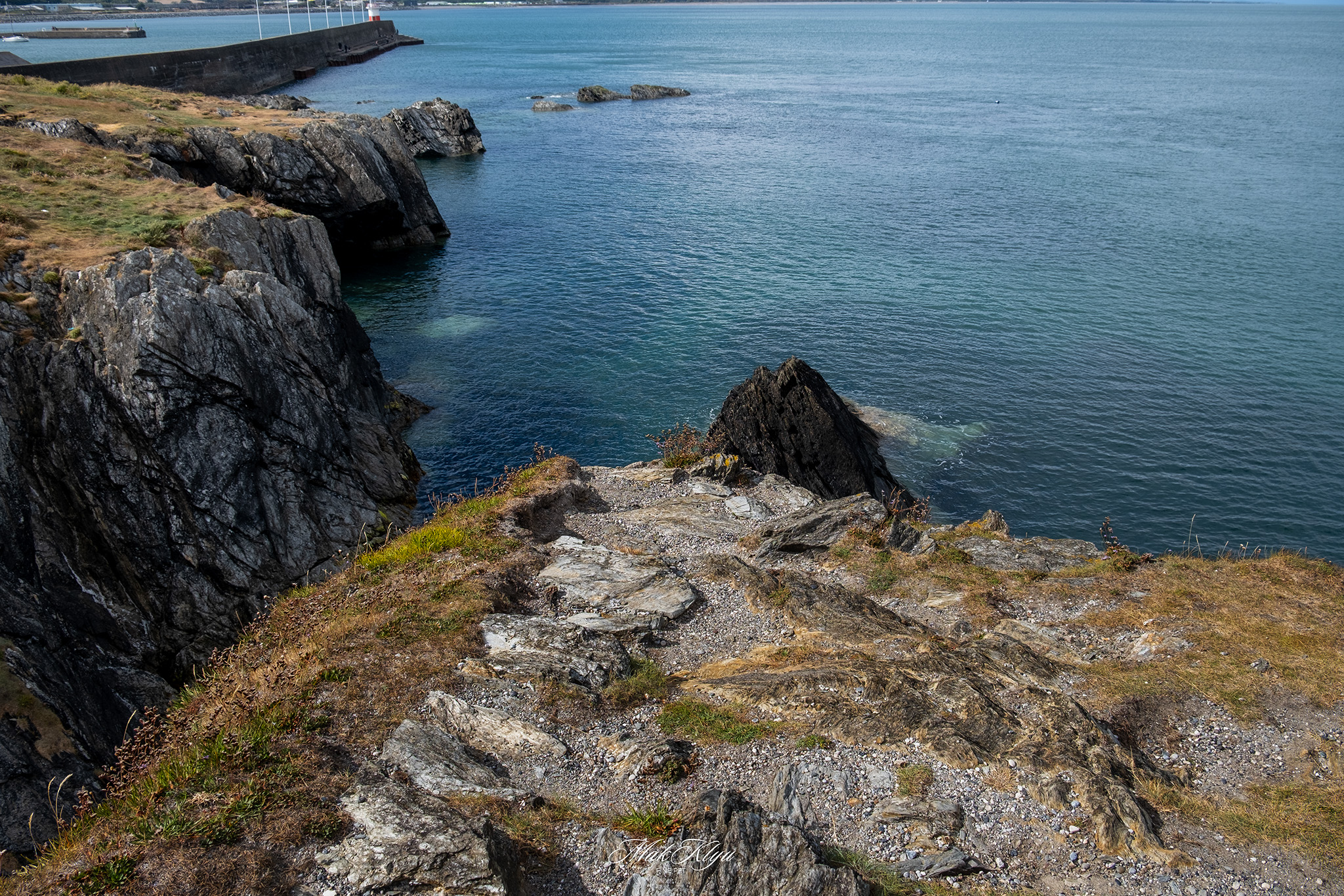 Rocky coastal cliffs descending into clear blue water beside a quiet harbour entrance